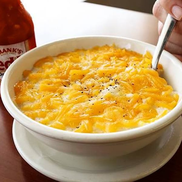 A bowl of cheesy grits with black pepper, a spoon, and a bottle of hot sauce.