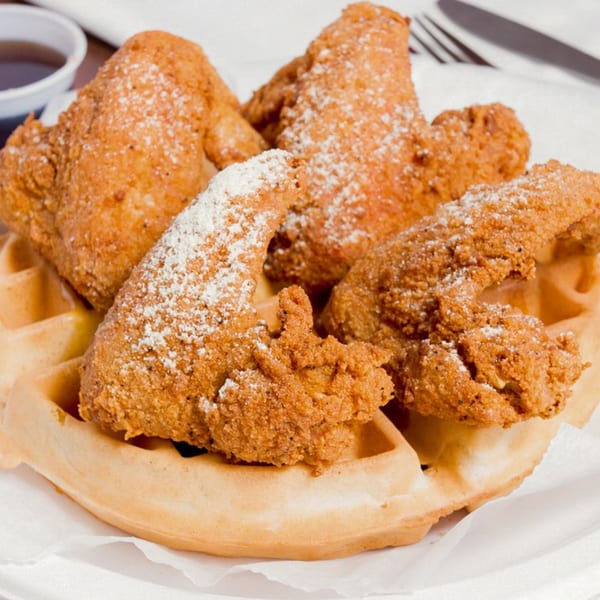 Four pieces of crispy fried chicken with powdered sugar on a waffle, with syrup in the background.