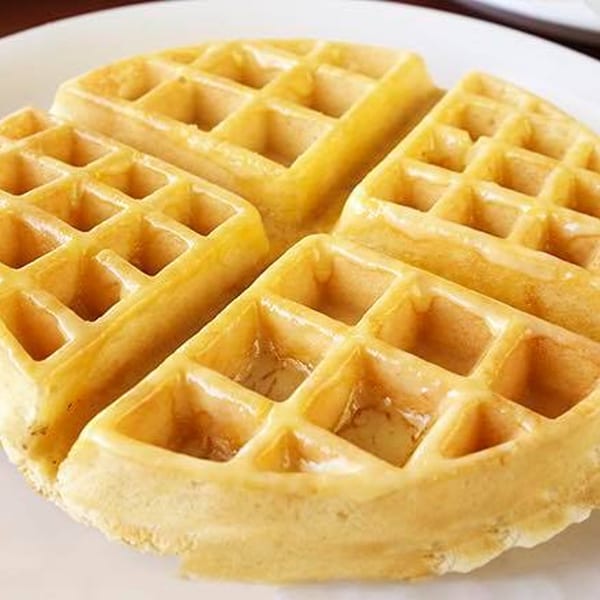 A golden-brown waffle, quartered, with syrup glistening in its square pockets, on a white plate.