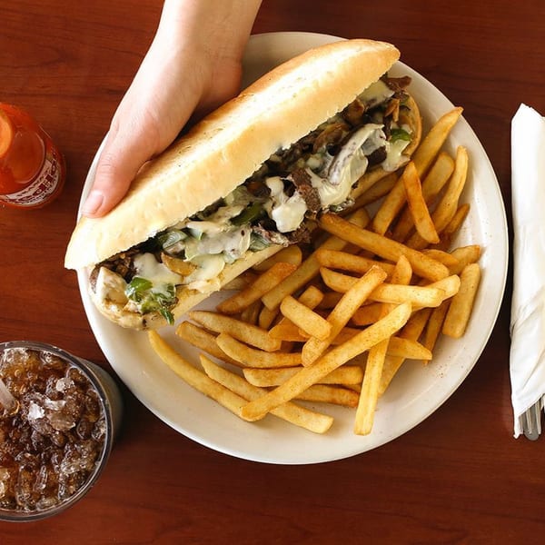 Hand holding a cheesesteak sandwich with fries, a drink, and hot sauce on a wooden table.